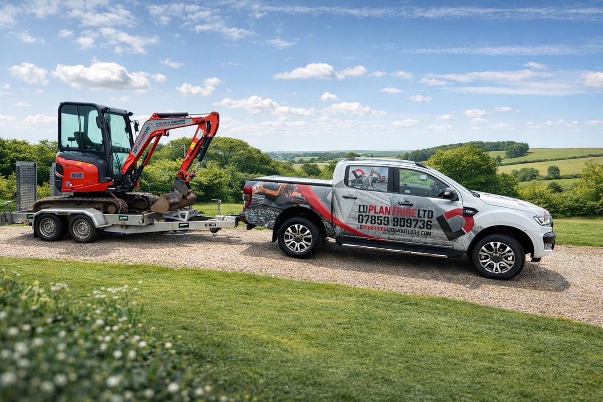 Red and gray mini excavator on a trailer towed by a white LD Plant Hire pickup truck in the Isle of Wight countryside